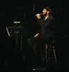 A bearded man in black clothing with his eyes closed. He is holding a microphone and sitting on a stool on a stage.