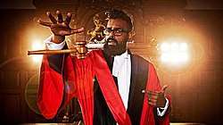 Romesh wears red robes in a courtroom as a gavel floats in the air