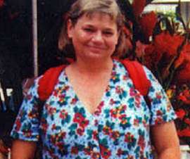 A middle-aged woman with light-colored hair in a light blue print top with the red straps of a backpack around her shoulders