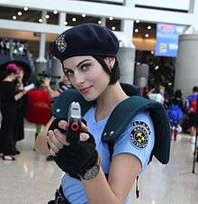 A woman wearing a costume at a convention from the waist up. She is wearing a dark blue beret, light blue shirt with shoulder armour and black fingerless gloves. She is pointing a pistol towards the viewer