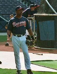 A dark-skinned man in a navy-blue baseball jersey and cap and white baseball pants standing on a grass field wearing a black baseball glove on his left hand