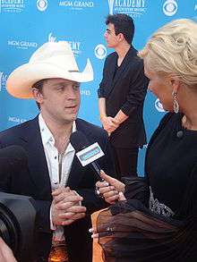 A young man in a dark jacket and white cowboy hat talking into a microphone being held by a blonde-haired woman.