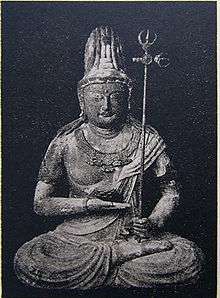 Front view of a statue in lotus position. The palm of the right hand is turned upward and held in front of the stomach. The left hand, close to the right foot, is holding a long pole with decorations at the end resembling a trident.