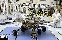 A robotic rover rests on a blue floor, while several scientists wearing white bodysuits observe it.