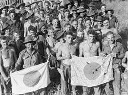 A crowd of men, some shirtless, some wearing slouch hats, hold up two rising sun flags. One has Japanese writing on it.