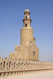 Photo of a minaret in four levels, with battlements in the foreground