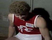 A footballer with curly blond hair in a red and white jersey looks down in front of a set of lockers