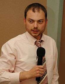 A balding, bearded Russian man wearing a pink shirt and striped necktie, holding a microphone, with a gold wedding ring on his finger.