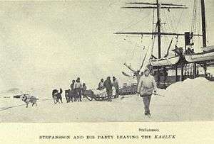 In the foreground a white-clad figure strides towards the camera. Behind him a group of men and dogs stand around a loaded sledge. To the right can be seen the upper parts of a ship, with heavy snow piled up at its sides.