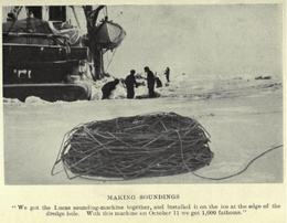 In the foreground, standing on sea ice, is a massive coil of rope or steel wire. In the background three men stand by a hole in the ice. Part of a ship's hull is visible, left background.