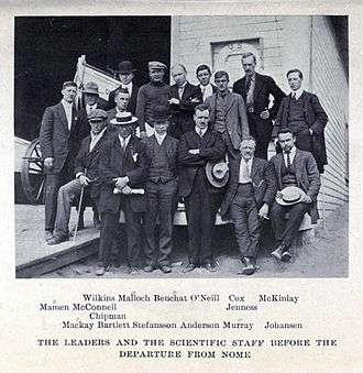 A group of 16 men, standing or sitting, on the deck of a ship with a small lifeboat visible, left background. The group's pose is casual and the men are variously attired, many in suits with casual hats of different sorts.