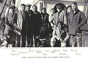 Ten men, one woman and two children stand (one man crouching) on a ship's deck. Both children are largely obscured in shadow. They are warmly dressed, mostly in thick jackets and boots, and the facial expressions of most are sombre and weary, although a few are attempting to smile.