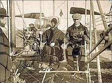 Wilbur and Katharine Wright seated in the Wright Model A Flyer with Orville Wright standing nearby. This was Katharine's first time flying. Her skirt is tied with a string.