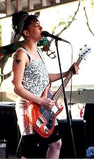 A female musician, Kathleen Hanna, singing into a microphone and playing electric bass. She is performing with the band she leads, Bikini Kill.