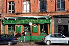 Green-fronted Irish pub, with "Katy Daly's" written on the hoarding above. A black-fronted door is to the right, and cars are parked on the roadway in front.