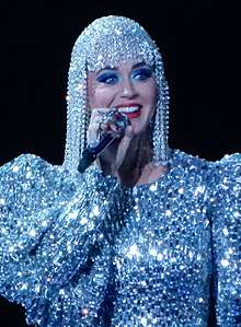 Katy Perry, a woman wearing a jeweled wig, smiling on stage wearing a jeweled outfit. She is holding a microphone to her mouth in her right hand.
