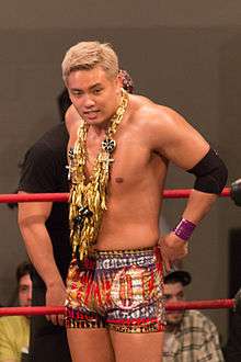 Kazuchika Okada, a Japanese man with blonde hair, wearing a golden garland as well as red and gold trunks while standing in a professional wrestling ring
