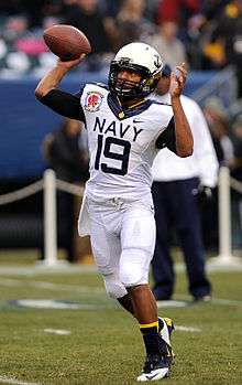 An American football player in a white uniform with "Navy" across the front throws a ball to a receiver out of frame.