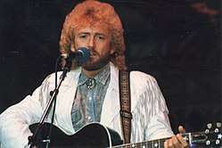 A man with long curly hair and a beard, wearing a white jacket and grey shirt, playing a guitar and singing into a microphone
