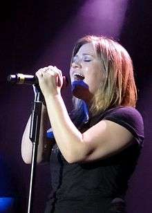 An image of an blonde haired woman, wearing a black T-shirt, singing against a dark violet background