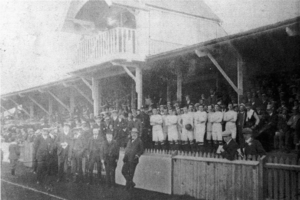 A faded black-and-white photograph of an early 20th-century football stand, crowded with people. An impressive canopy is built into the stand's roof.