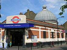 A red-bricked building with a rectangular, dark blue sign reading "KENNINGTON STATION" in white letters all under a light blue sky