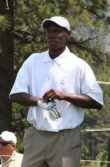 A dark-skinned man wearing a white polo shirt, white crownless baseball cap, and a white golf glove, which he is in the process of adjusting