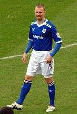 A man with a shaven head wearing a blue football jersey, white shorts and blue socks. He is standing on a grass pitch