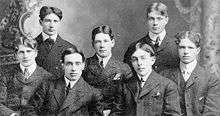 Seven men apparently in their 20s pose for a half-length group photo. They are wearing formal suits and ties.