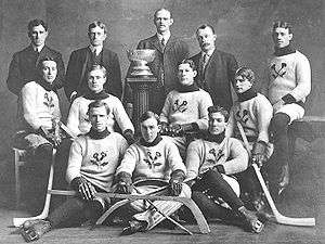 An early ice hockey team poses for a photo. Eight players, all seated around a trophy on a pedestal, are dressed in wool sweaters with a thistle emblem. They wear skates and hold ice hockey sticks. Behind them stand four men in suits.