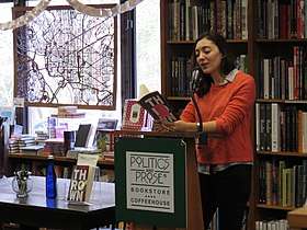 A photograph of Kerry Howley. She is standing at a podium with a Politics and Prose sign, in front of a large shelf of books. In the background is a window with a carved wooden map of the District of Columbia hanging in it. Howley has her eyes turned down, reading from her book. She is wearing an orange sweater with the sleeves pushed up and a grey collar shirt underneath.