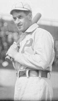 A man in an old-style white baseball uniform with a large block "P" over the left breast and crownless cap holds a baseball bat over his right shoulder.