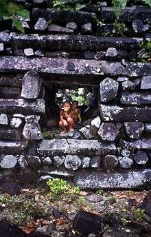 Image of two children crouching in a square passage through a wall constructed of dark grey columnar basalt pieces.