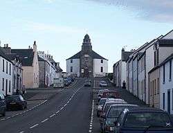 A wide street leads up a slope with parked cars and stone houses painted in whites and yellows on either side. At the end of the street there is a grey and white building with a short spire.