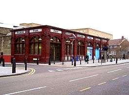 A red-bricked building with a series of white signs together reading "KILBURN PARK STATION" in red letters and people walking in front all under a white sky