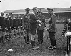 A black and white photo of a rugby field in which three men in military uniform, one of whom is King George, present a silver trophy to a rugby player dressed in black kit. Behind in a line are the rest of the team.