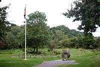 A clearing sparsely surrounded by trees and bushes. A gravel-lined spot is at the centre, sporting a stone with flowers lain in front of it. On the left stands a flagpole, whose flag lies unfurled.