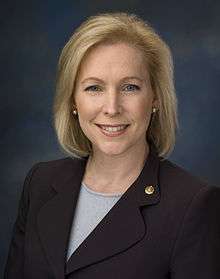A portrait shot of a smiling, middle-aged female looking straight ahead. She has chin-length blonde hair, and is wearing a dark blazer with a light top. She is placed in front of a nondescript background.