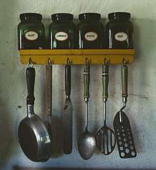 Various kitchen utensils.  At top: a spice rack with jars of mint, caraway, thyme, and sage.  Lower: hanging from hooks; a small pan, a meat fork, an icing spatula, a whole spoon, a slotted spoon, and a perforated spatula.