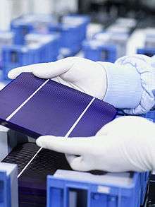 A part of gloved hands are hold a tablet-sized  solar cell with three panels.