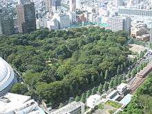 Bird eye view of a wooded park surrounded by tall buildings and adjacent to a rail line.