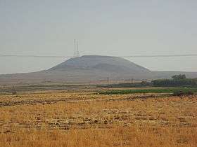 A black mountain with a telecommunication tower above it, seen from afar, and distorted by dusty mist. You are standing in a field overgrown by dry grasses, some of them are green.