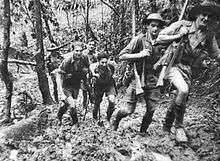 Soldiers in short sleeve shirts and shorts, slouch hats and helmets march up a muddy track carrying rifles slung over their shoulders