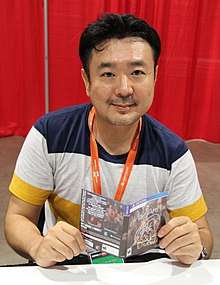 A 2016 photograph of Kotaro Uchikoshi, holding an autographed Zero Time Dilemma game cover