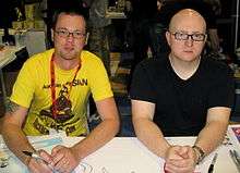 Two young men, Mike Krahulik and Jerry Holkins, sitting at a booth at a convention.
