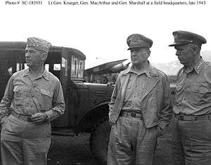 Three men in Army uniforms with open neck shirts. Krueger wears a garrison cap, MacArthur his special cap, and Marshall, a peaked cap. in the background there is a truck and a propeller-driven airplane.