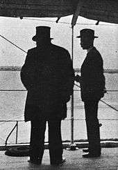 Kruger viewed in silhouette from behind, Bredell to his right. Kruger is wearing his top hat.