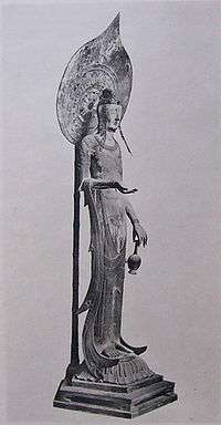 Three-quarter view of a very slim and tall statue carrying a vase in with two fingers of her left hand. Her right arm is bend with the palm of her right hand facing upward. A halo on a pole is seen behind the statue.