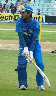 A man wearing a dark blue cap, not clean shaven, wearing a blue shirt and blue trousers, holding a cricket bat.