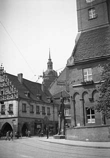 Black and white photograph of Kurfürstenhaus to the left and the New Town Hall to the right, at Hauptstrasse street in Brandenburg an der Havel. In the background is the tower of St. Katharinen church. A statue of knight Roland at the corner of the Town Hall.